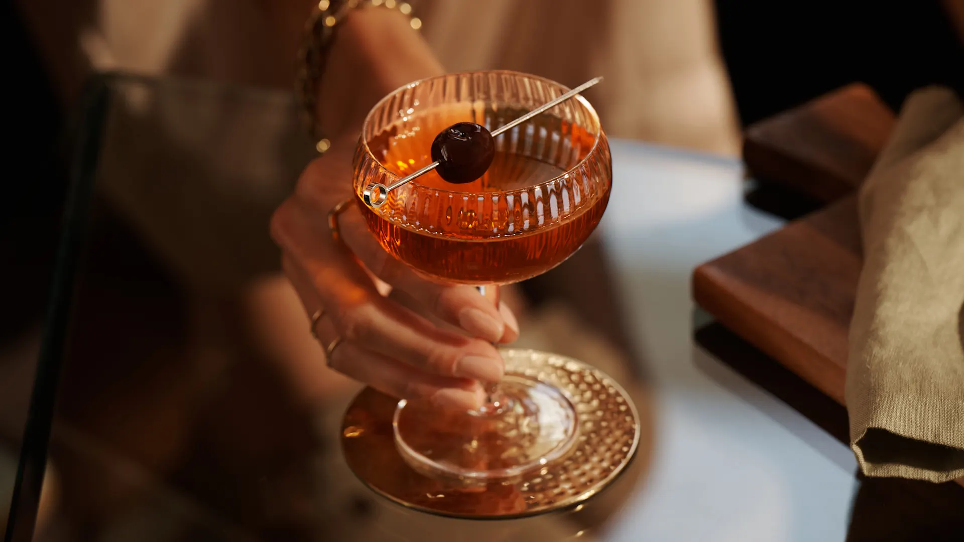 A Manhattan cocktail sits on a glass table, with a skewed maraschino cherry sitting over the rim of the coupe glass. A person holds the coupe by the stem.