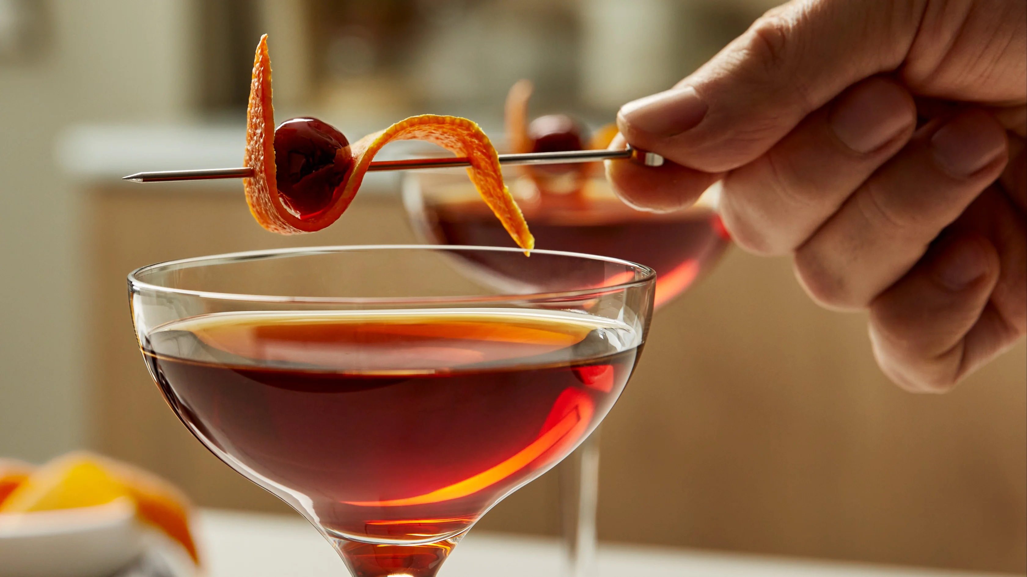 Eine Person legt eine aufgespießte Orangenschale und eine Kirsche auf den Rand eines braunen Cocktails in einem Coupé-Glas