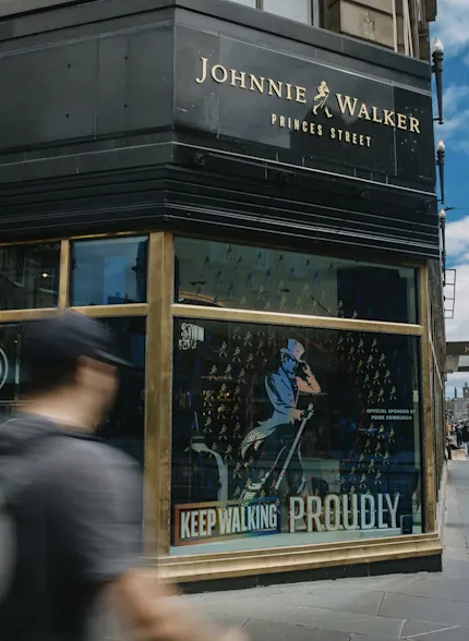 A Johnnie Walker employee stands on a stage, talking to a group of people seated in front. The employee gestures to the screen behind them which shows a projection of several wooden barrels.