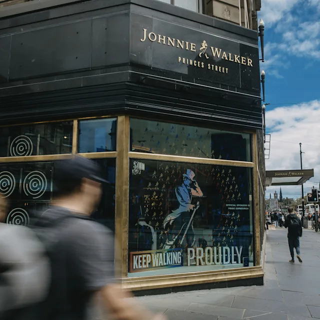 A Johnnie Walker employee stands on a stage, talking to a group of people seated in front. The employee gestures to the screen behind them which shows a projection of several wooden barrels.