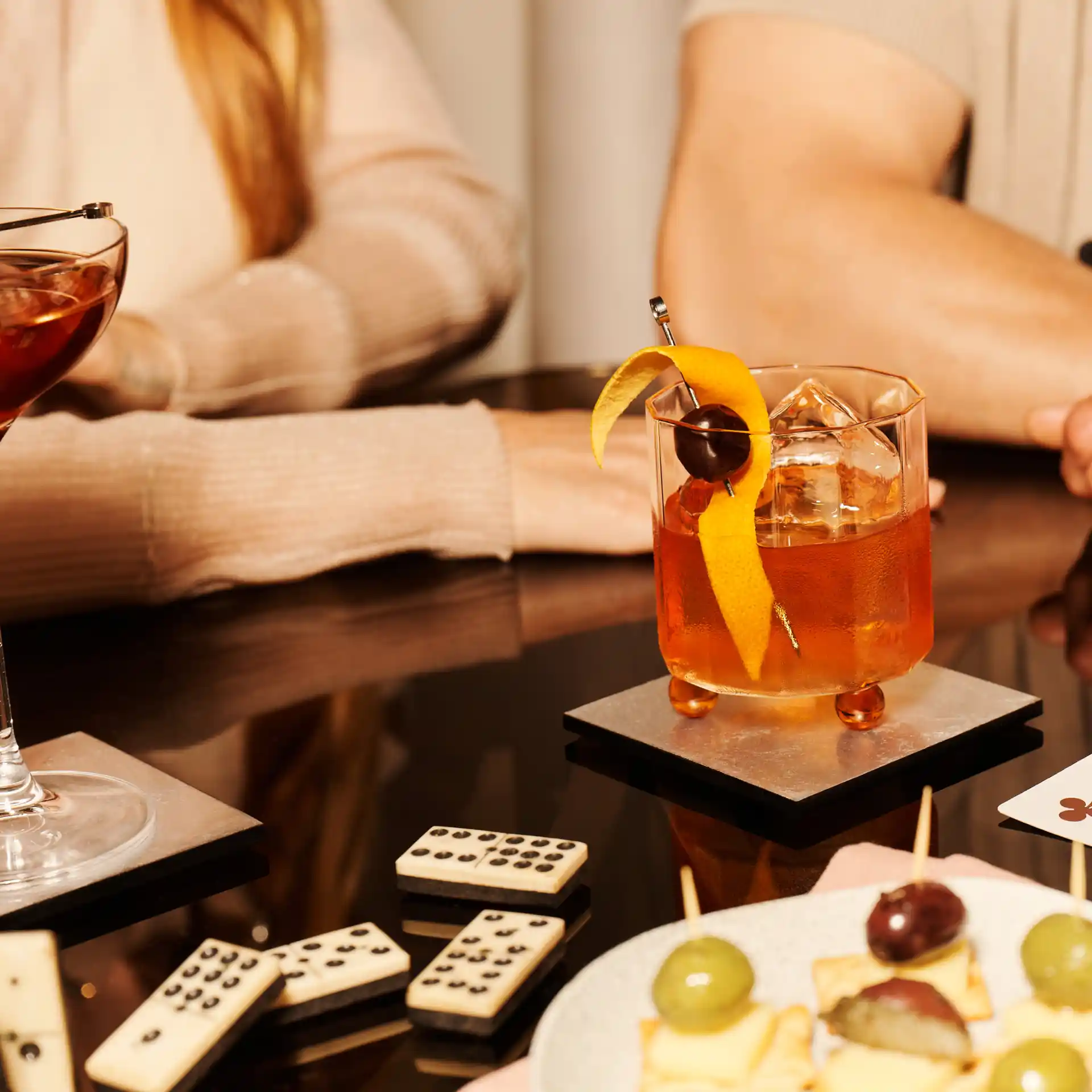 Ein Old-Fashioned-Cocktail steht auf einem Tisch, der von Dominosteinen und Spielkarten umgeben ist.