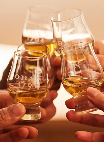 Four whisky tasting glasses containing whisky are held together in a cheers gesture. They are branded with Oban 1794.
