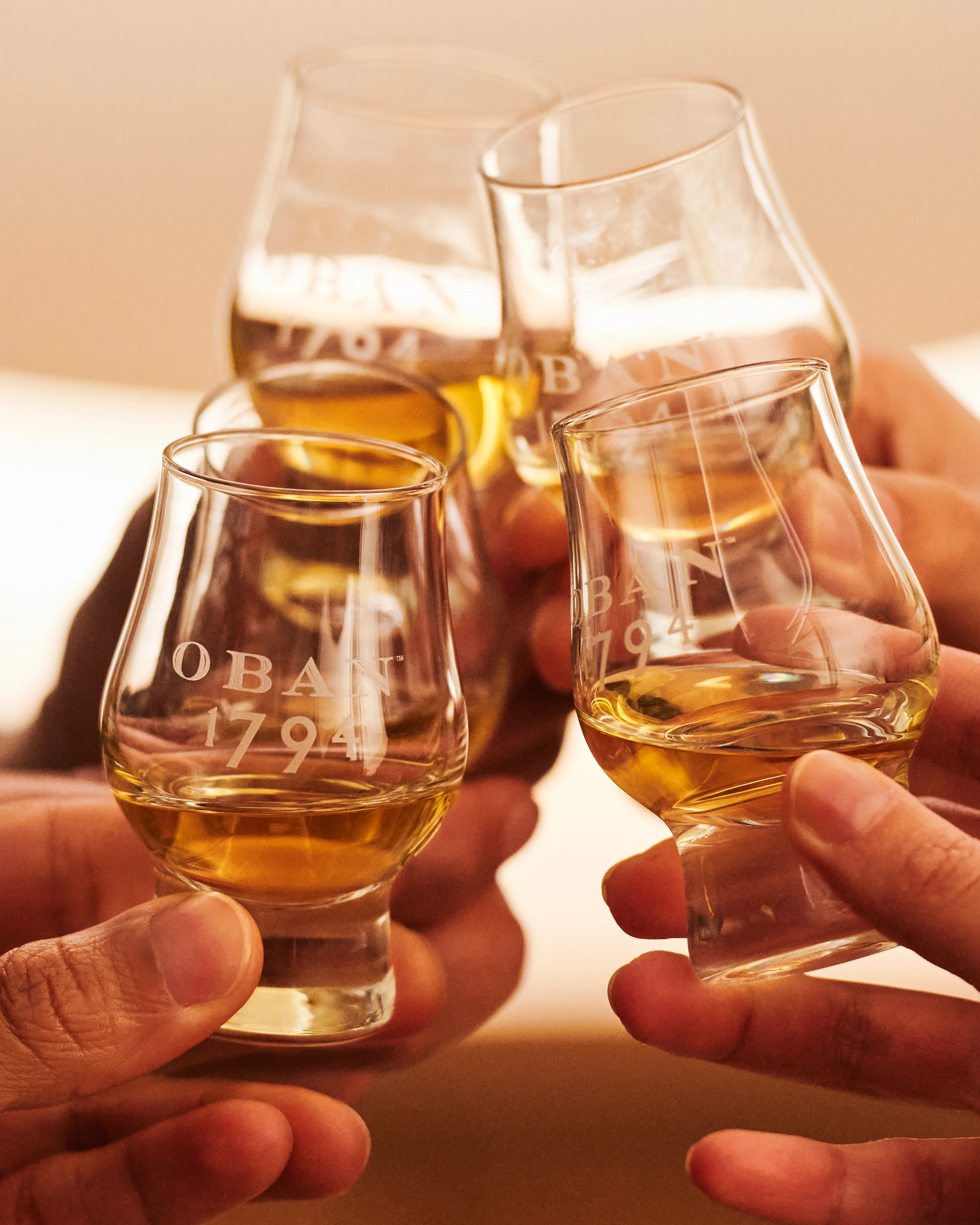 Four whisky tasting glasses containing whisky are held together in a cheers gesture. They are branded with Oban 1794.