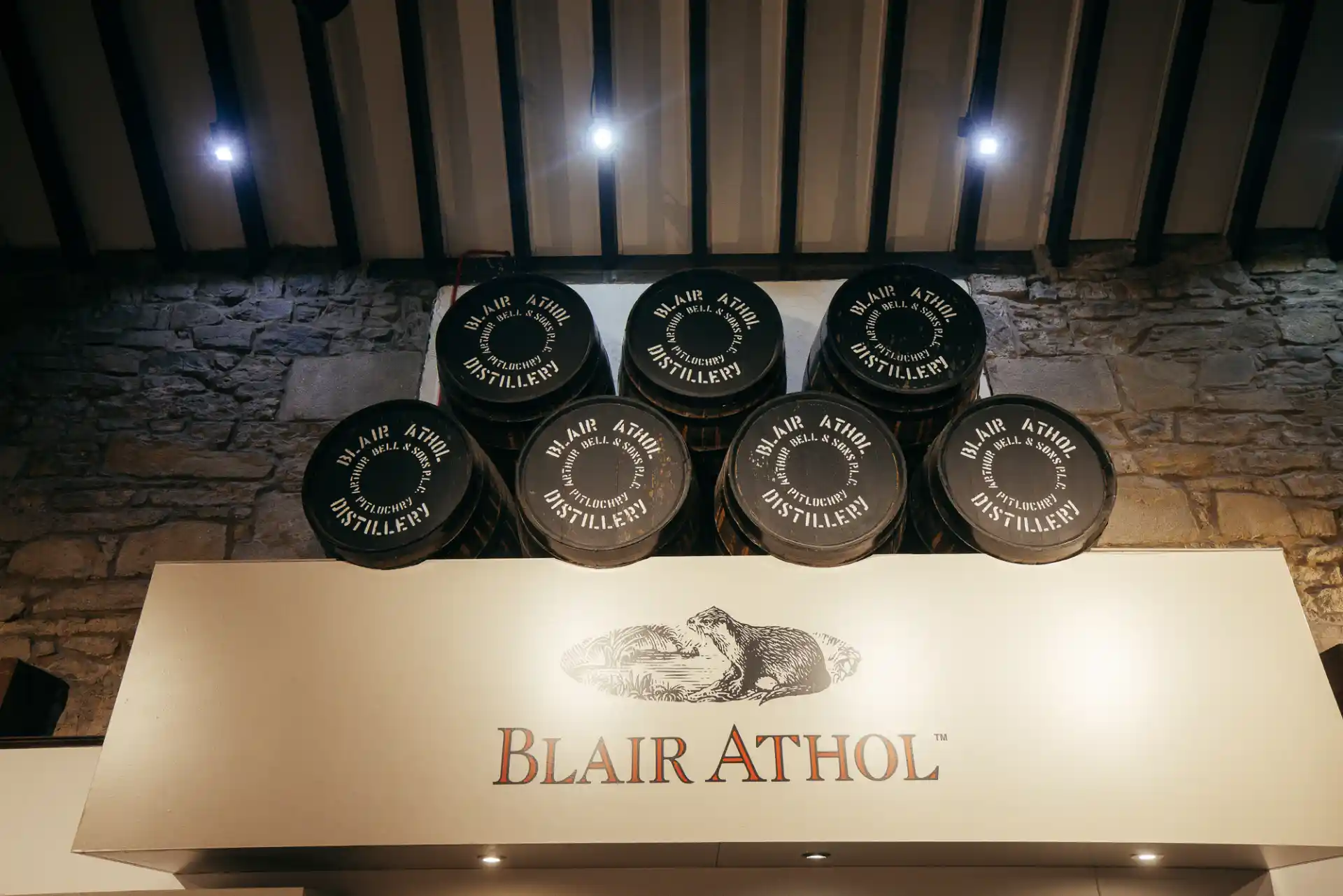 Zeven zwarte houten vaten met het opschrift 'Blair Athol Distillery' staan op elkaar gestapeld op een licht houten display met het Blair Athol logo.