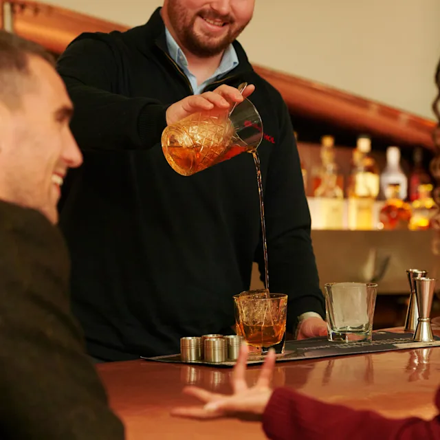 Two friends sit at the bar in Blair Athol distillery, watching a bartender pour out a whisky cocktail