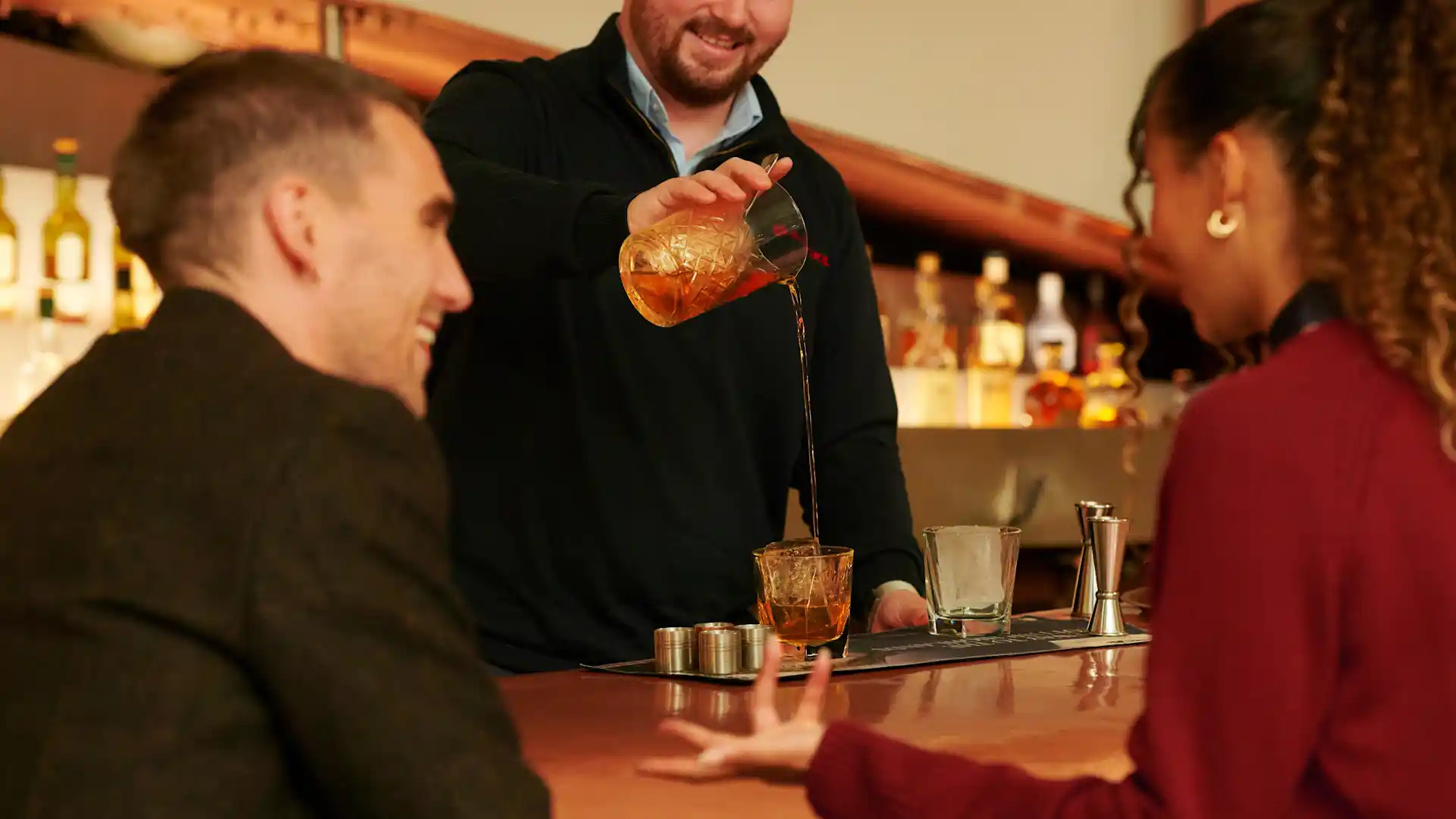 Two friends sit at the bar in Blair Athol distillery, watching a bartender pour out a whisky cocktail