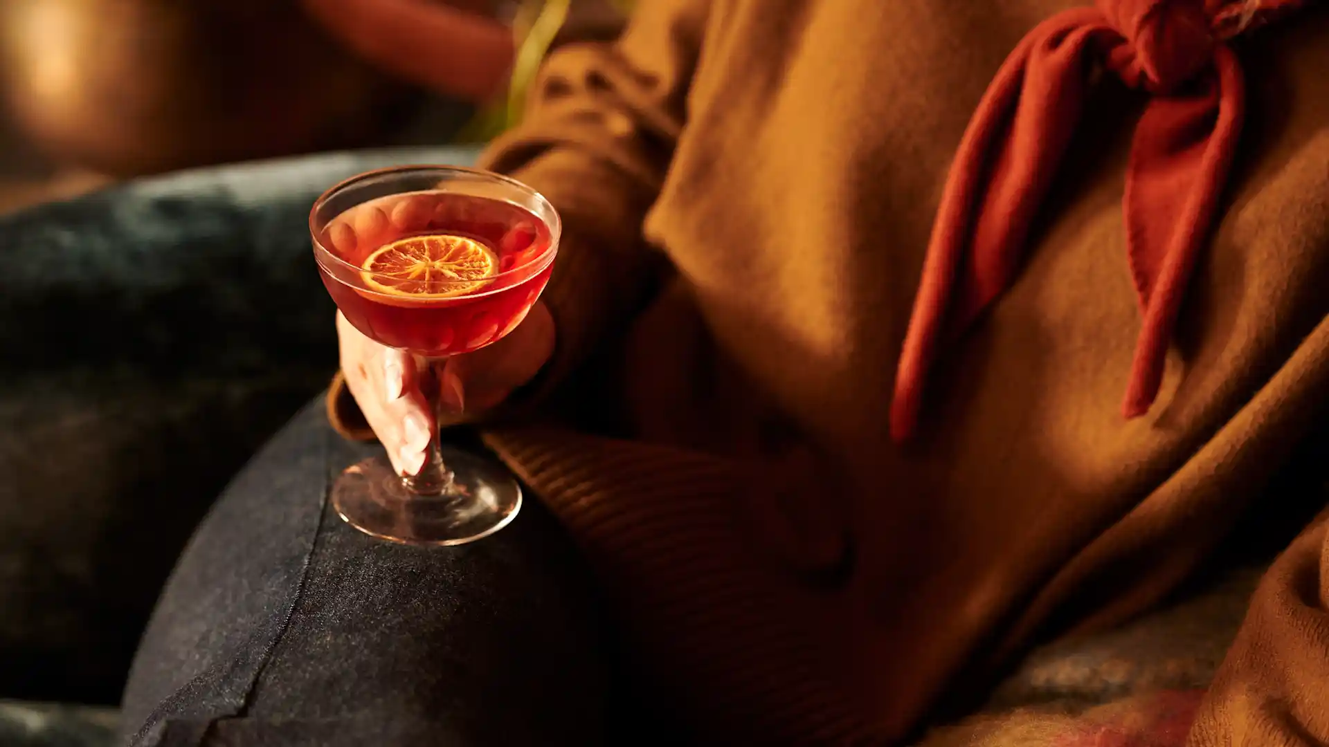 A woman holds a red-coloured whisky cocktail on her lap. The cocktail is in a coupe glass and is garnished with a slice of dried orange.