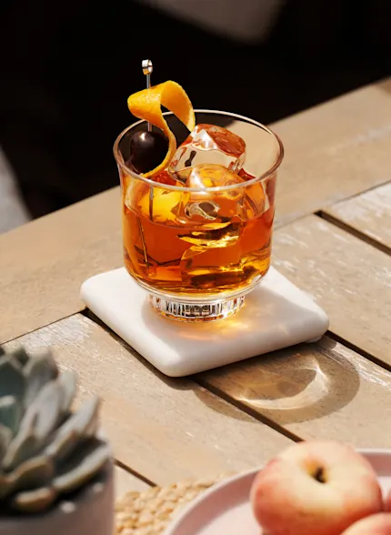 An Old Fashioned cocktail sits on a wooden surface, surrounded by peaches and a small plant.