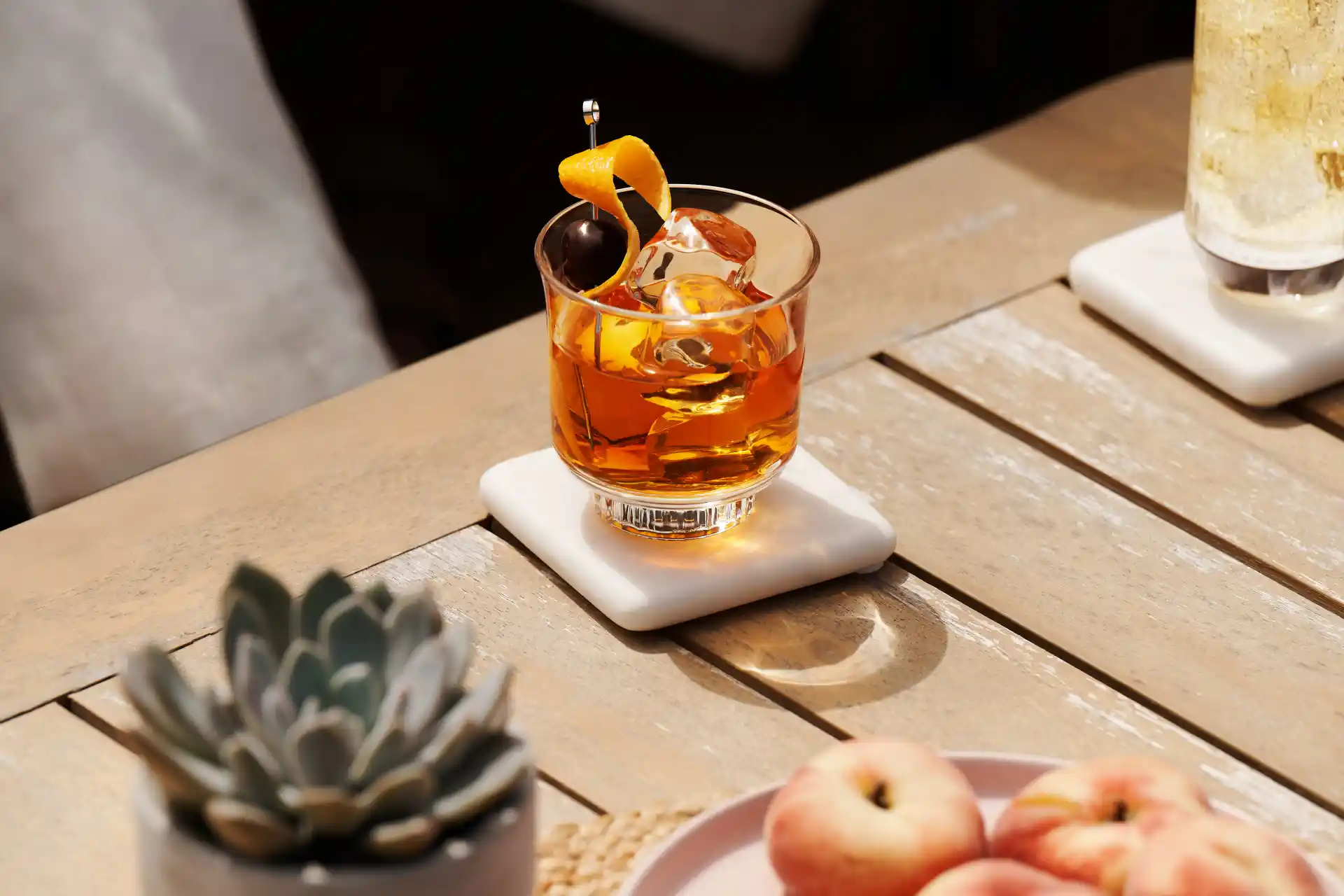 An Old Fashioned cocktail sits on a wooden table surrounded by a bowl of peaches, a plant and a highball cocktail