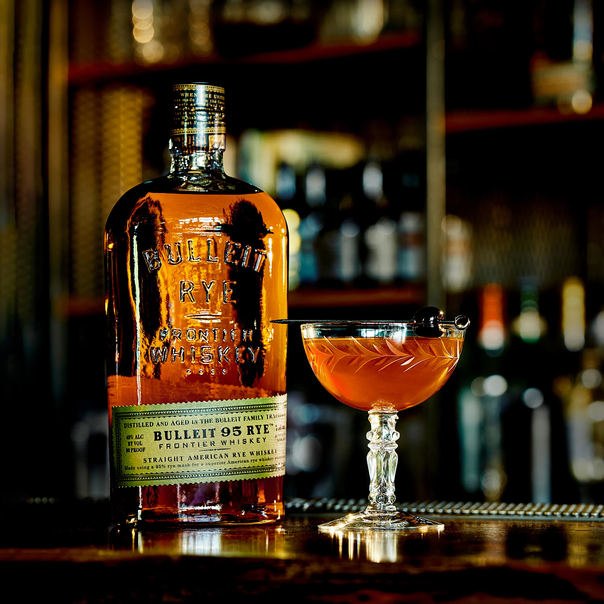 A bottle of Bulleit rye whiskey sits on a bar top next to a Manhattan cocktail in a coupe glass with a skewered cherry laying across the rim