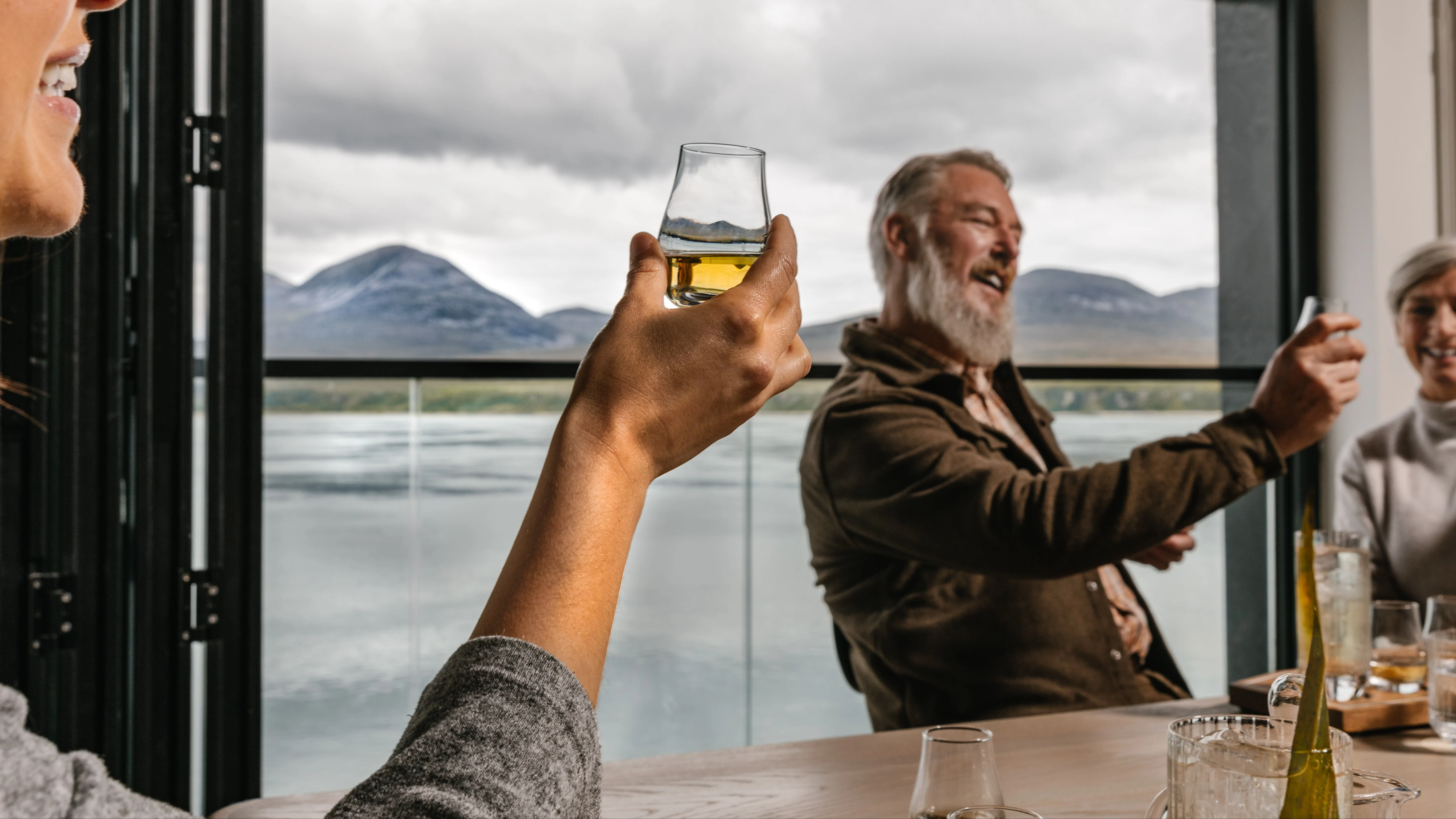 A person holds a glass of whisky up to the light around a table of friends, near a Scottish lake.