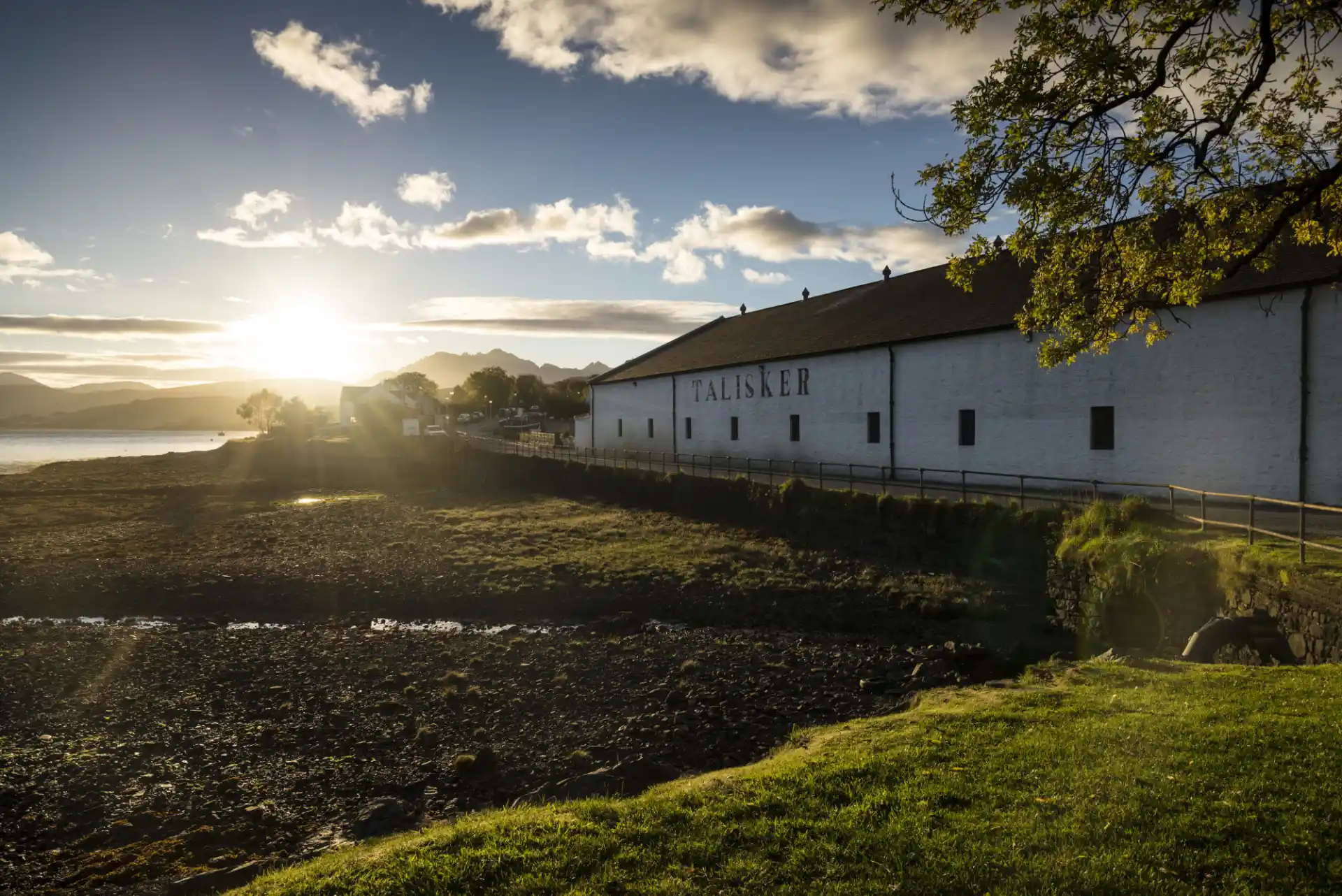 Talisker distillery sits on a pebbly shore on a green verge. It has white walls and the words Talisker are painted on the side in black.