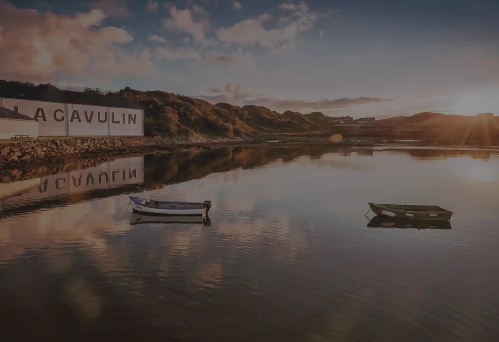 Two small boats sit on a body of water in front of a white building with the words Lagavulin painted onto the wall. Next to the building are grass-covered hills; the sky is blue and dotted with white clouds.