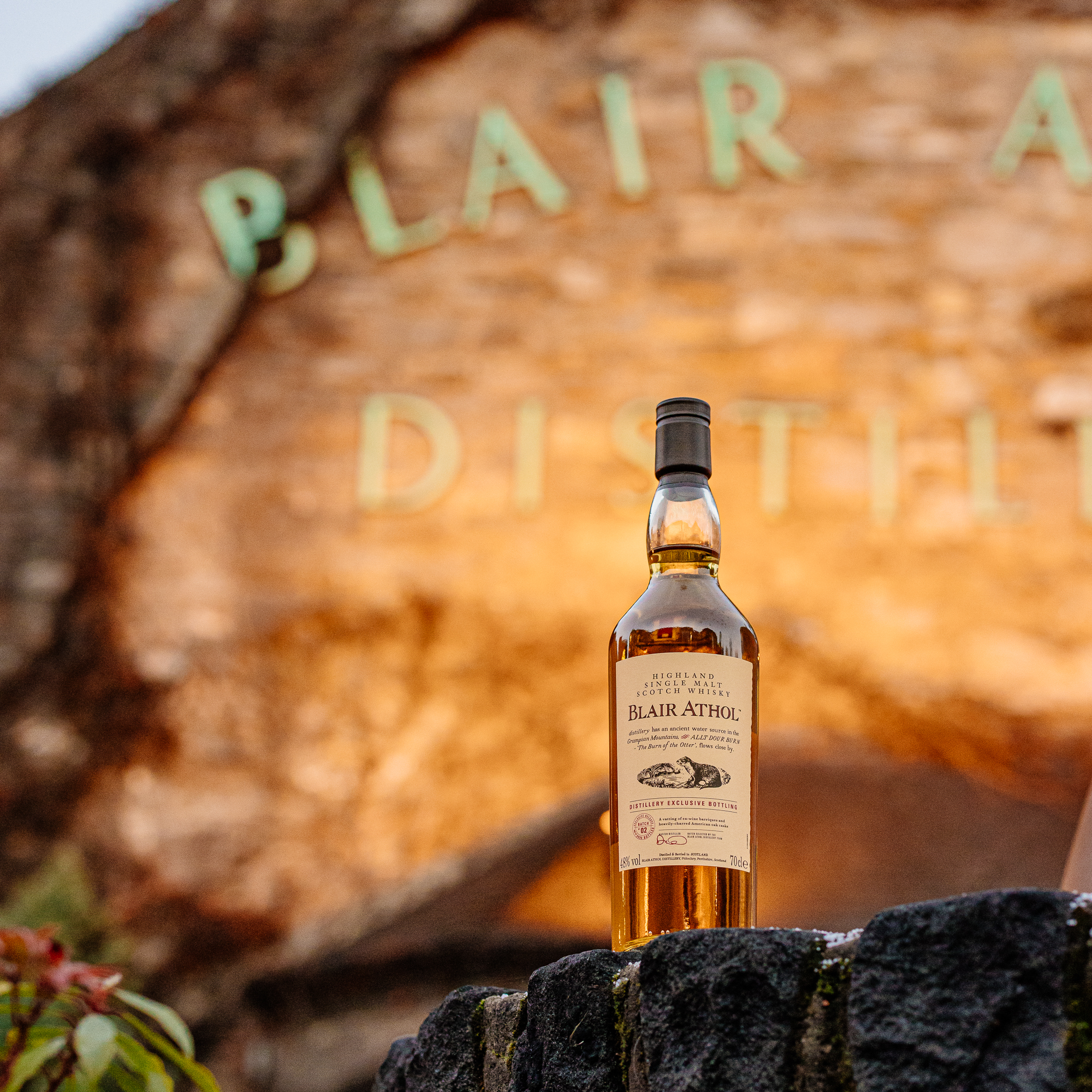 A bottle of Blair Athol whisky sits in front of the distillery sign which is lit with a golden light