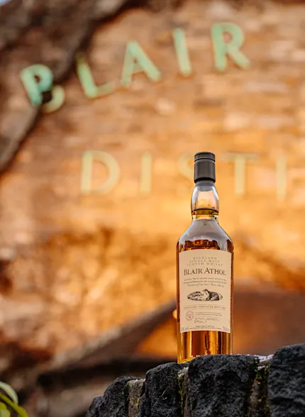 A bottle of Blair Athol whisky sits in front of the distillery sign which is lit with a golden light