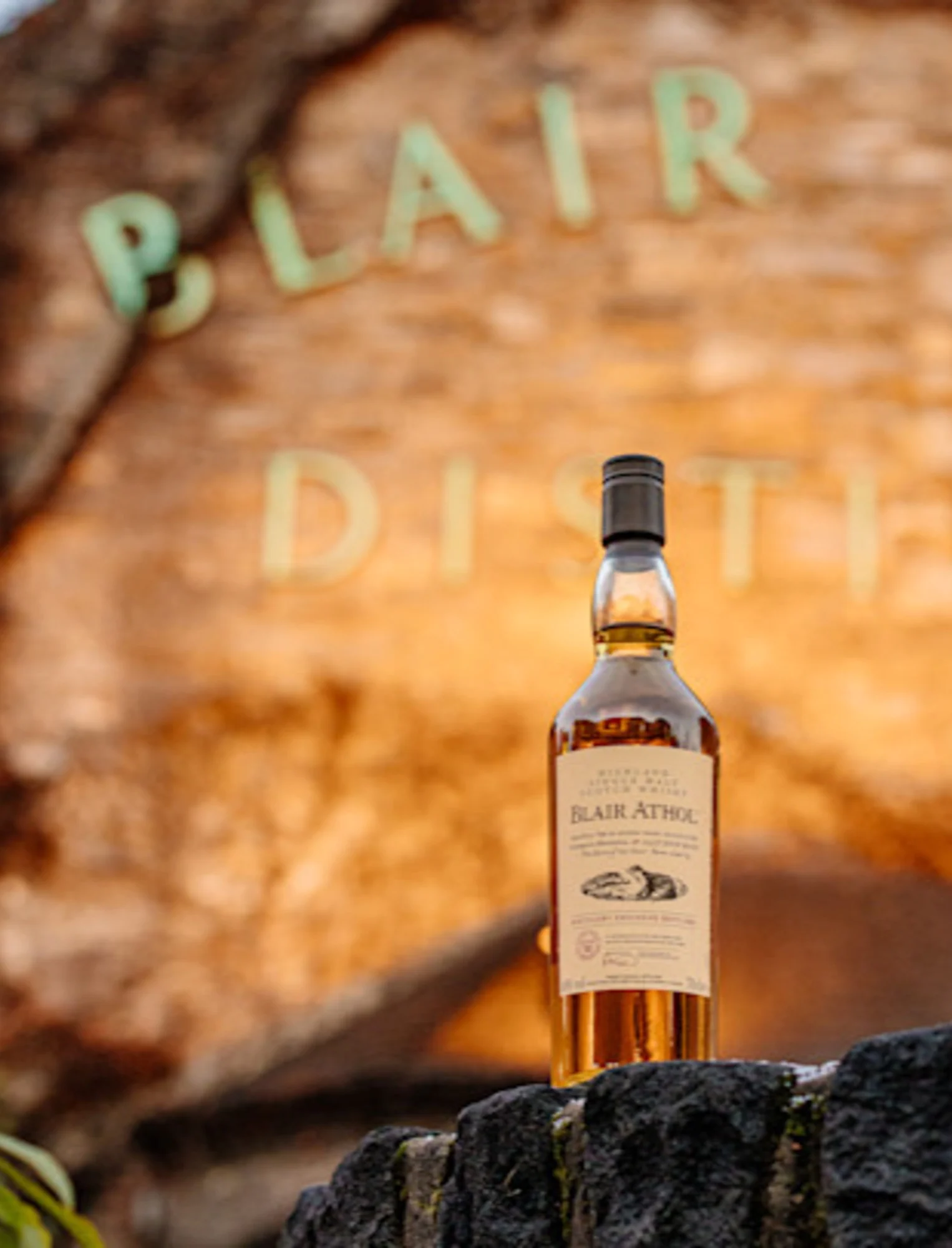 A bottle of Blair Athol whisky sits in front of the distillery sign which is lit with a golden light