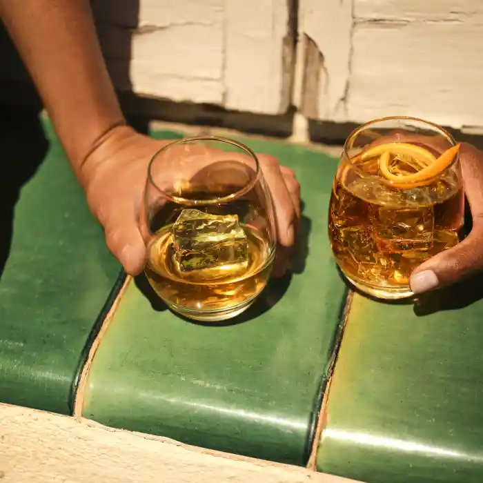 Two glasses of whisky sit on a green tile, both glasses are held by hands