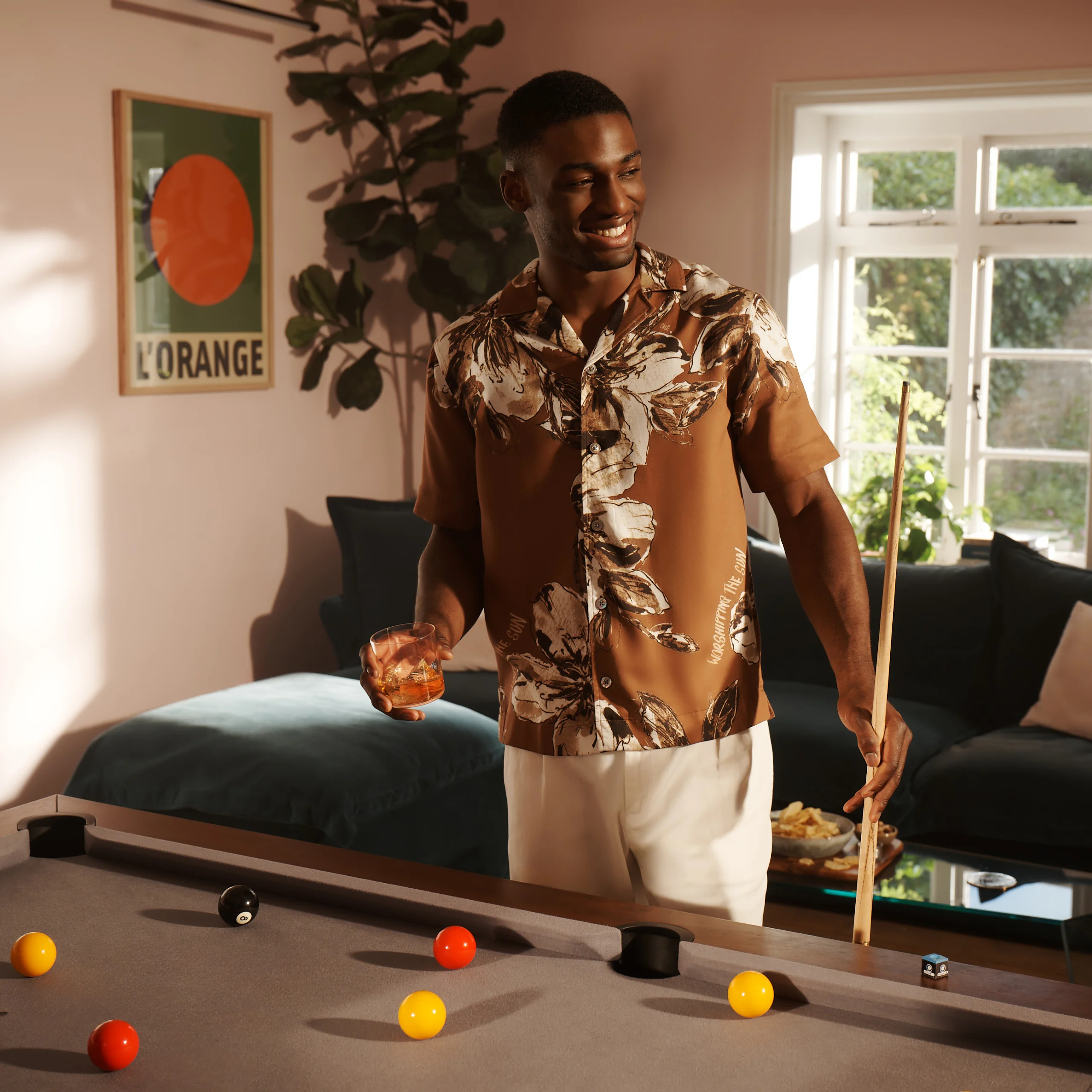 A man stands over a pool table, holding a pool cue in one hand and a rocks glass with whisky in the other. He is smiling.