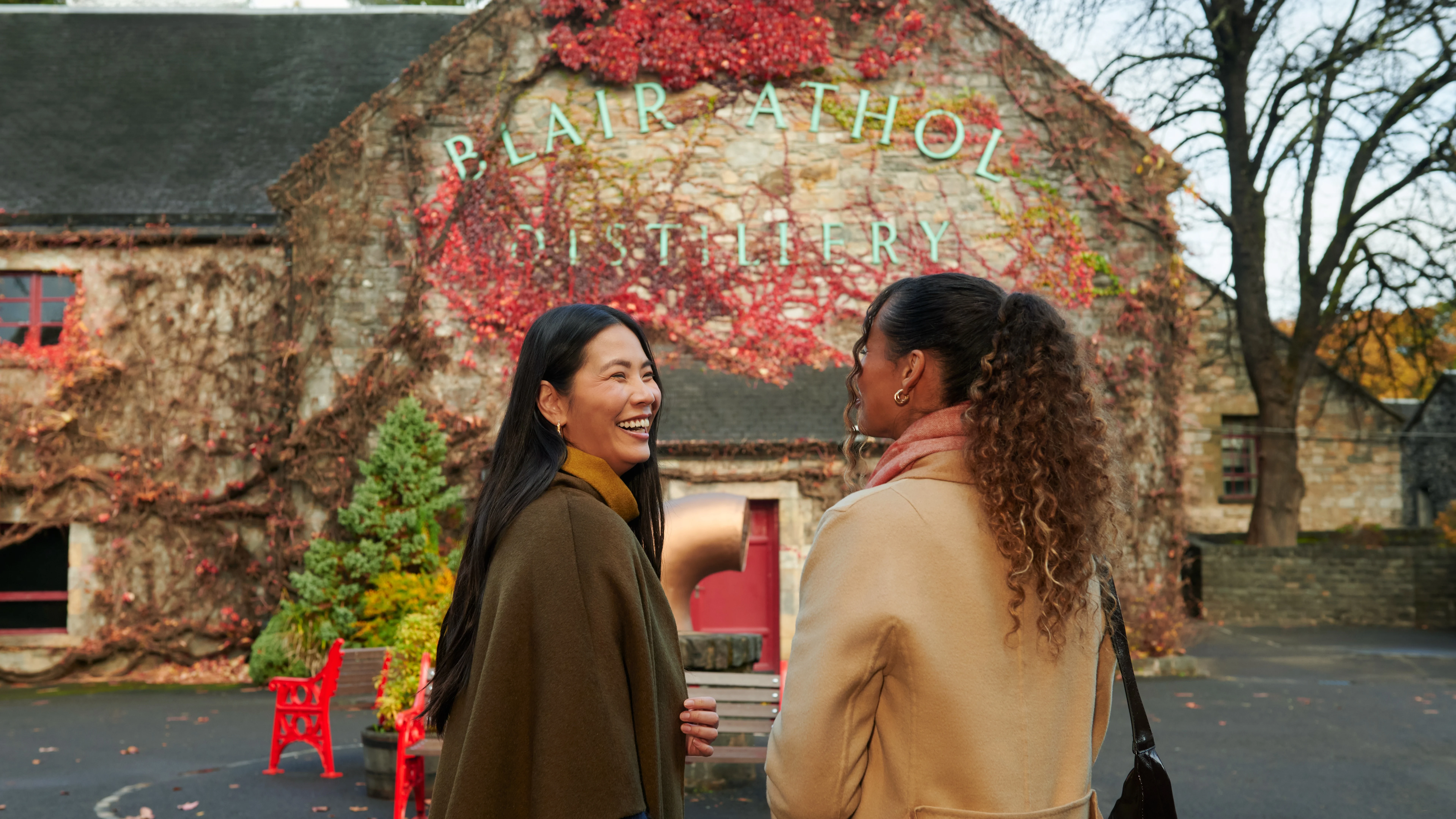 Two female friends stand outside Blair Athol distillery laughing and smiling