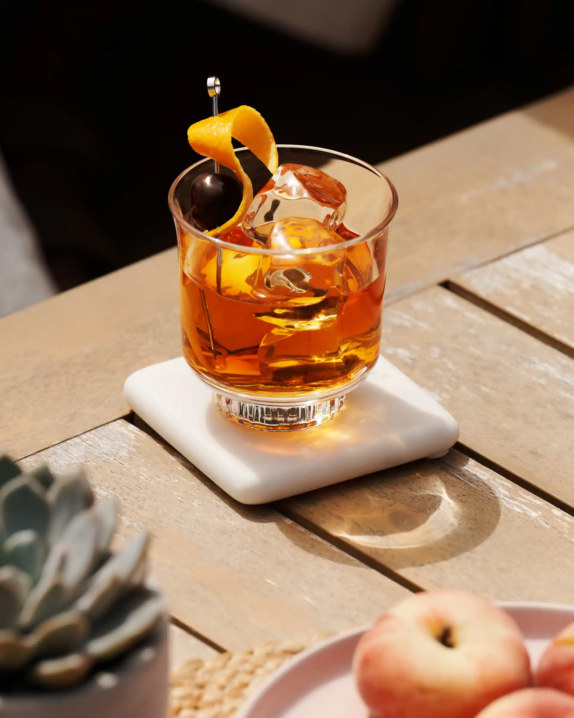 An Old Fashioned cocktail sits on a wooden table surrounded by a bowl of peaches, a plant and a highball cocktail