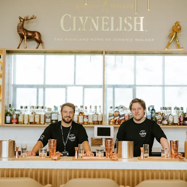 Two smiling Clynelish employees behind a bar. shelves display assorted whisky bottles, sign saying ‘Clynelish’. On the bar are assorted cocktail shakers and glasses; in front of the bar are three chairs.