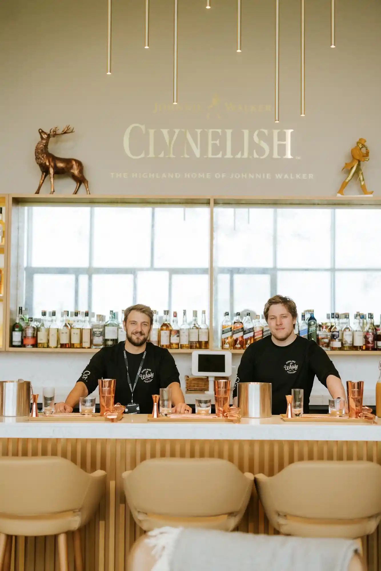 Twee lachende Clynelish-medewerkers achter een bar. Op planken staan verschillende whiskyflessen, op het bord staat 'Clynelish'. Op de bar staan diverse cocktailshakers en glazen; voor de bar staan drie stoelen.