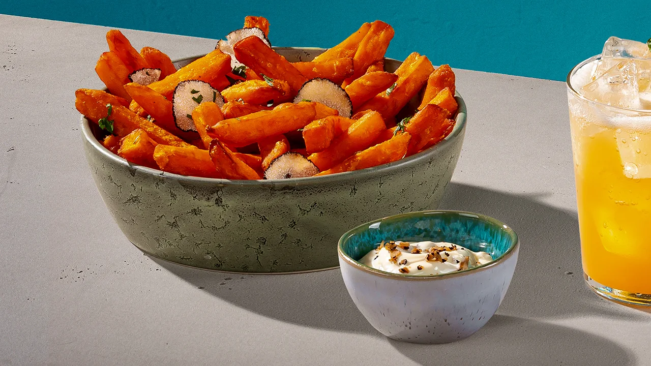 A glass of yellow whisky cocktail next to a bowl of sweet potato fries and a smaller bowl of truffle mayo.