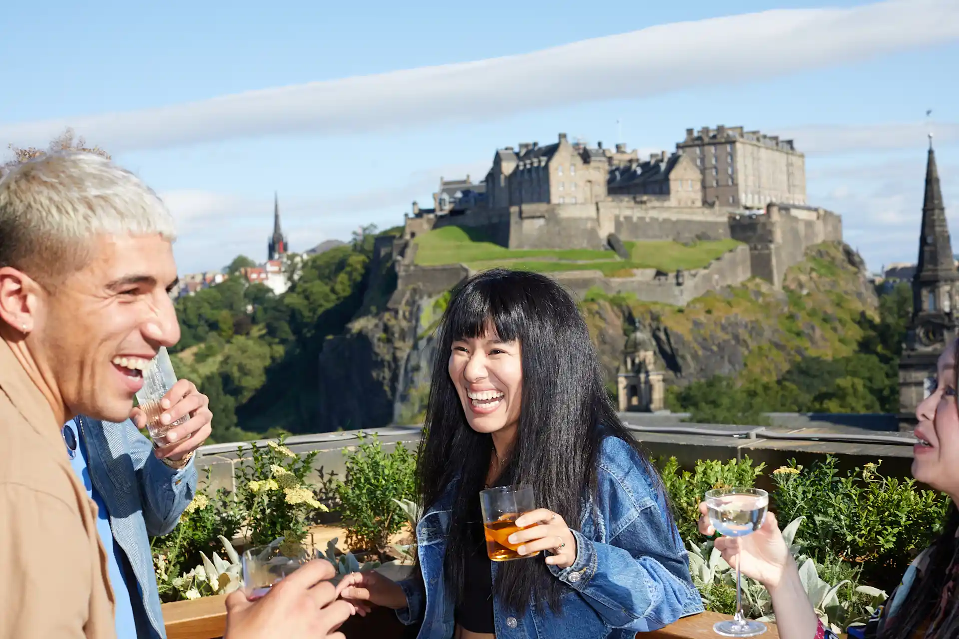 Eine Gruppe von Freunden genießt im Sommer Cocktails auf einer Dachterrassenbar mit Blick auf die Burg von Edinburgh