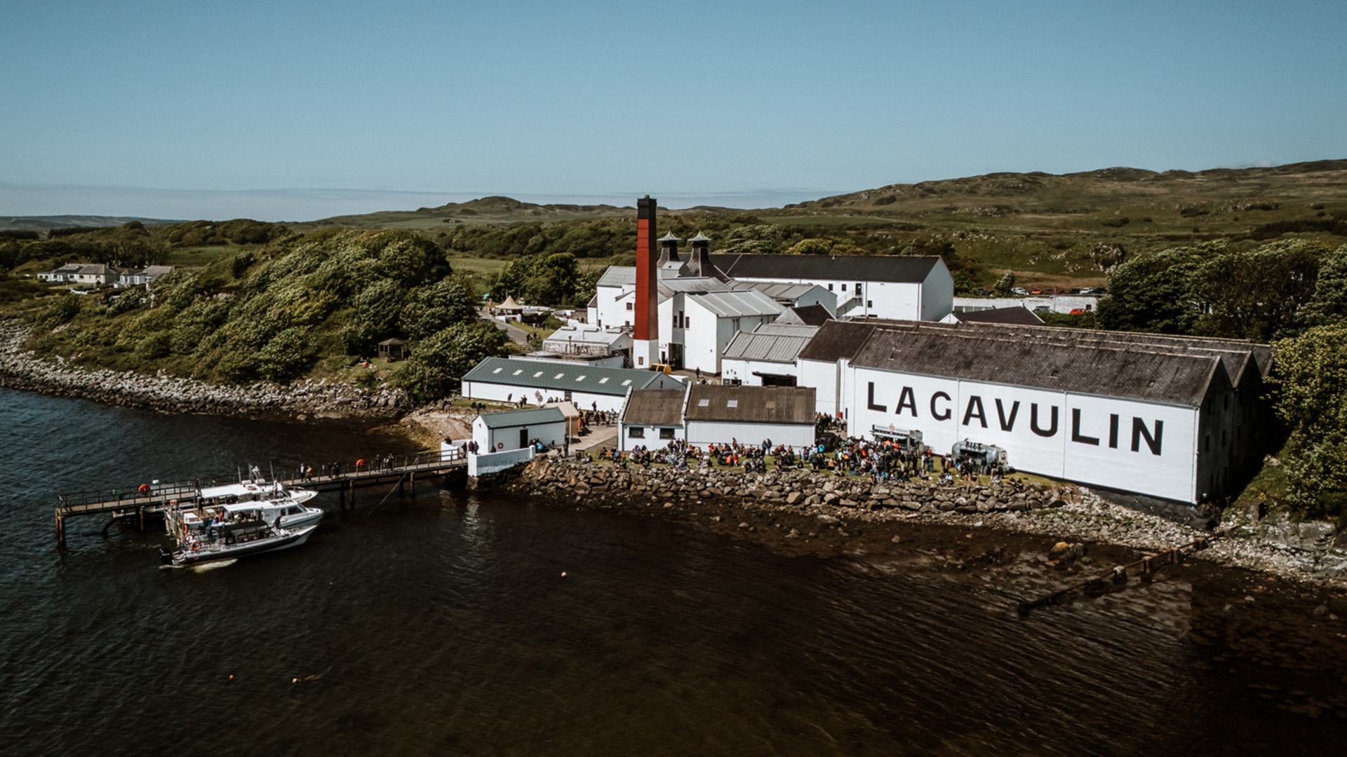 Eine Luftaufnahme der Lagavulin Destillerie auf Islay, Schottland. Das weiße Gebäude mit dem großen "LAGAVULIN" Schriftzug steht direkt an der Küste. Eine Menschenmenge versammelt sich vor der Destillerie, während Boote am Pier anlegen.