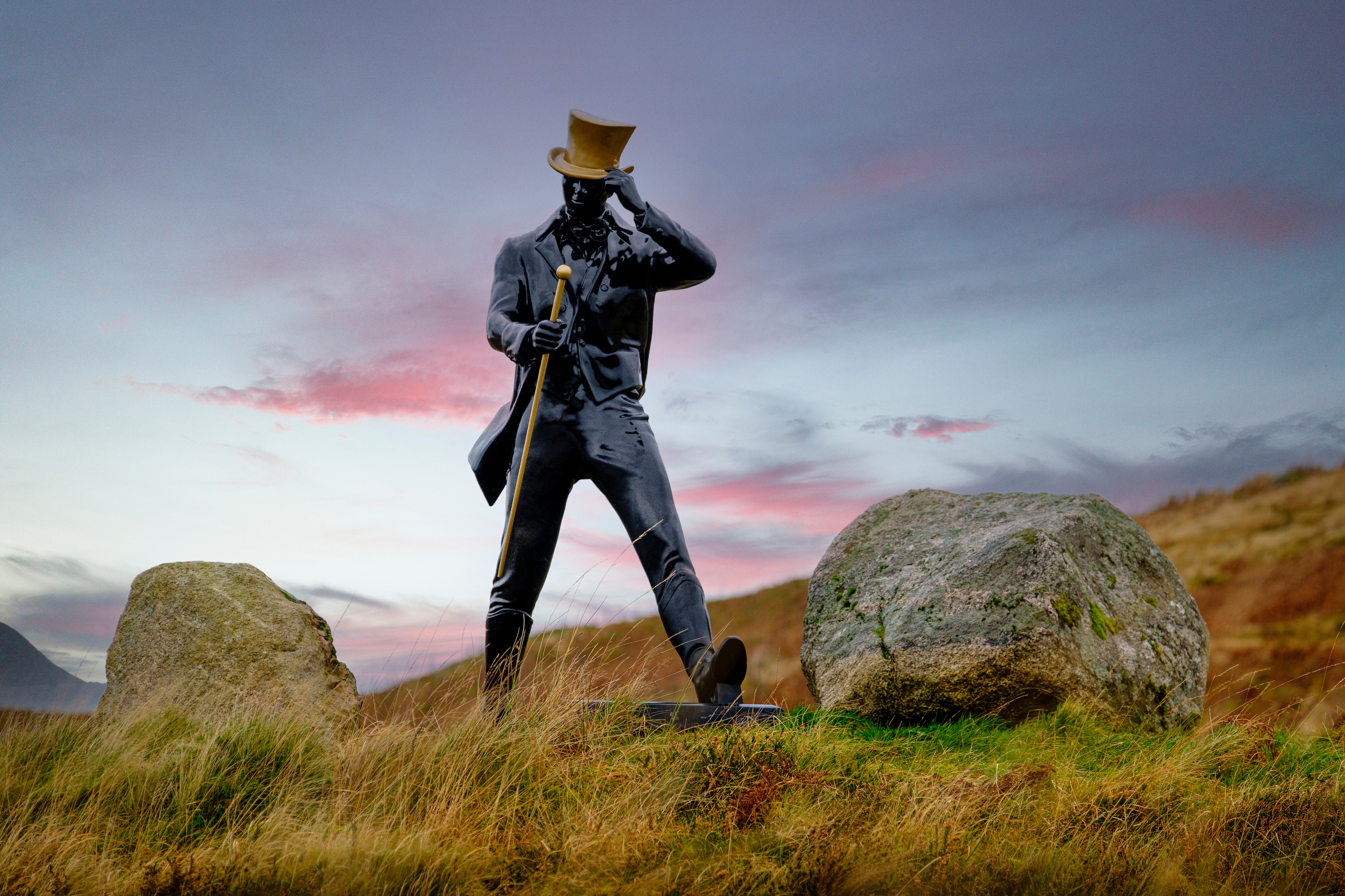 A statue of the Johnnie Walker ‘Striding Man’ stands on a grassy hillside flanked by two large rocks. The statue has a golden walking cane and top hat. Behind the statue is a blue sky with clouds.