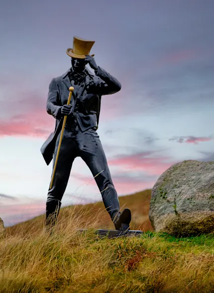 A statue of the Johnnie Walker ‘Striding Man’ stands on a grassy hillside flanked by two large rocks. The statue has a golden walking cane and top hat. Behind the statue is a blue sky with clouds.