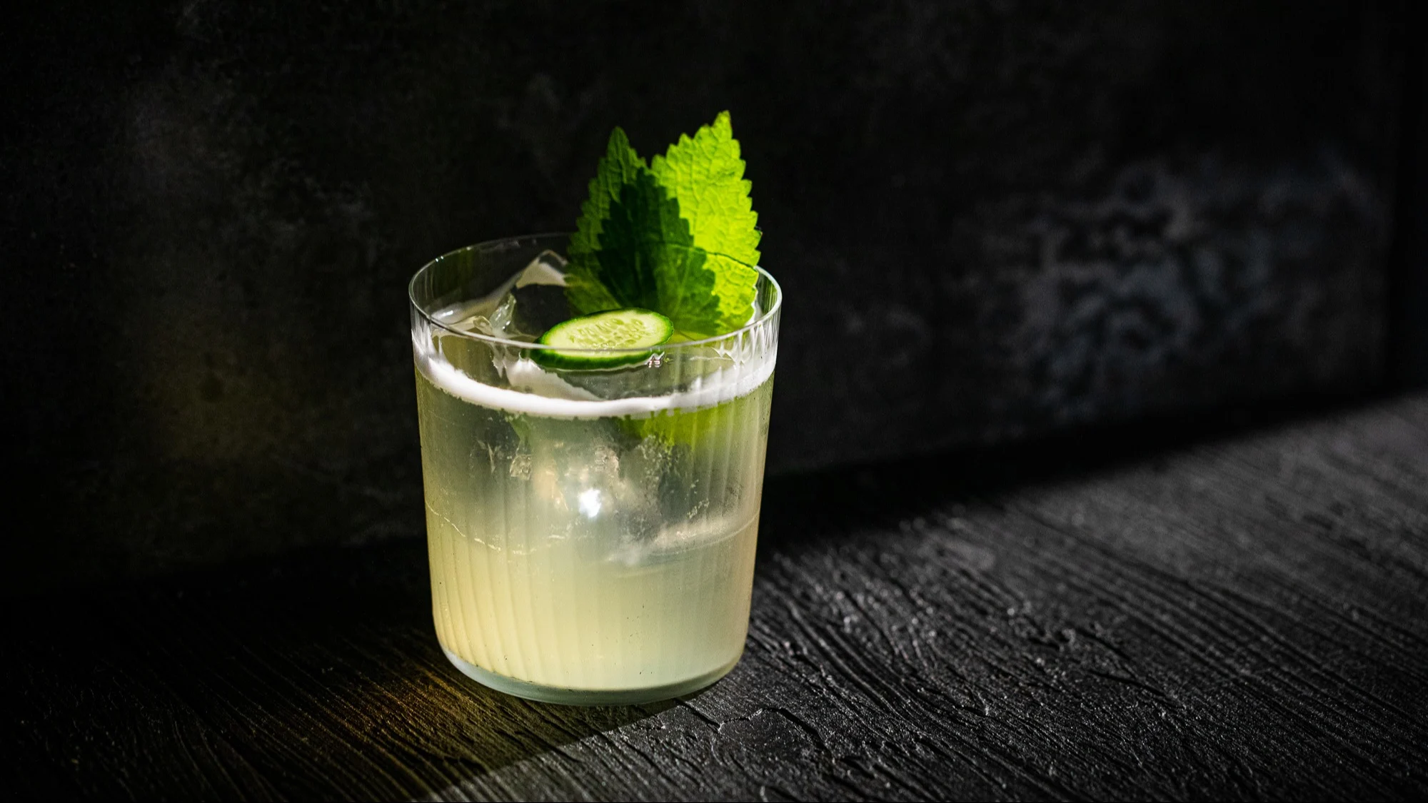 A pale cocktail in a rocks glass, with ice, a cucumber slice and two mint leaves