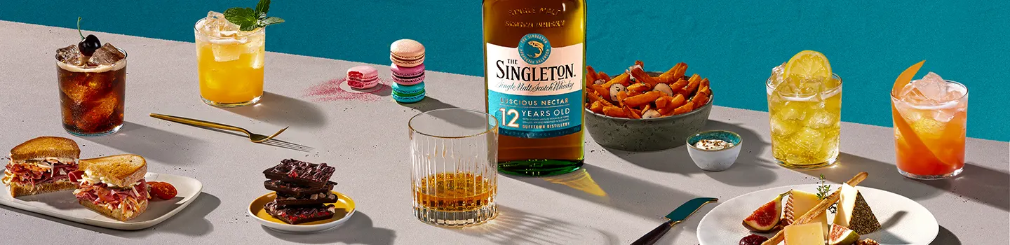 An assortment of food and drinks on a table with a bottle of The Singleton 12 Year Old Single Malt Scotch Whisky. The spread includes cocktails, a sandwich, macarons, chocolate, fries, cheese, and figs.