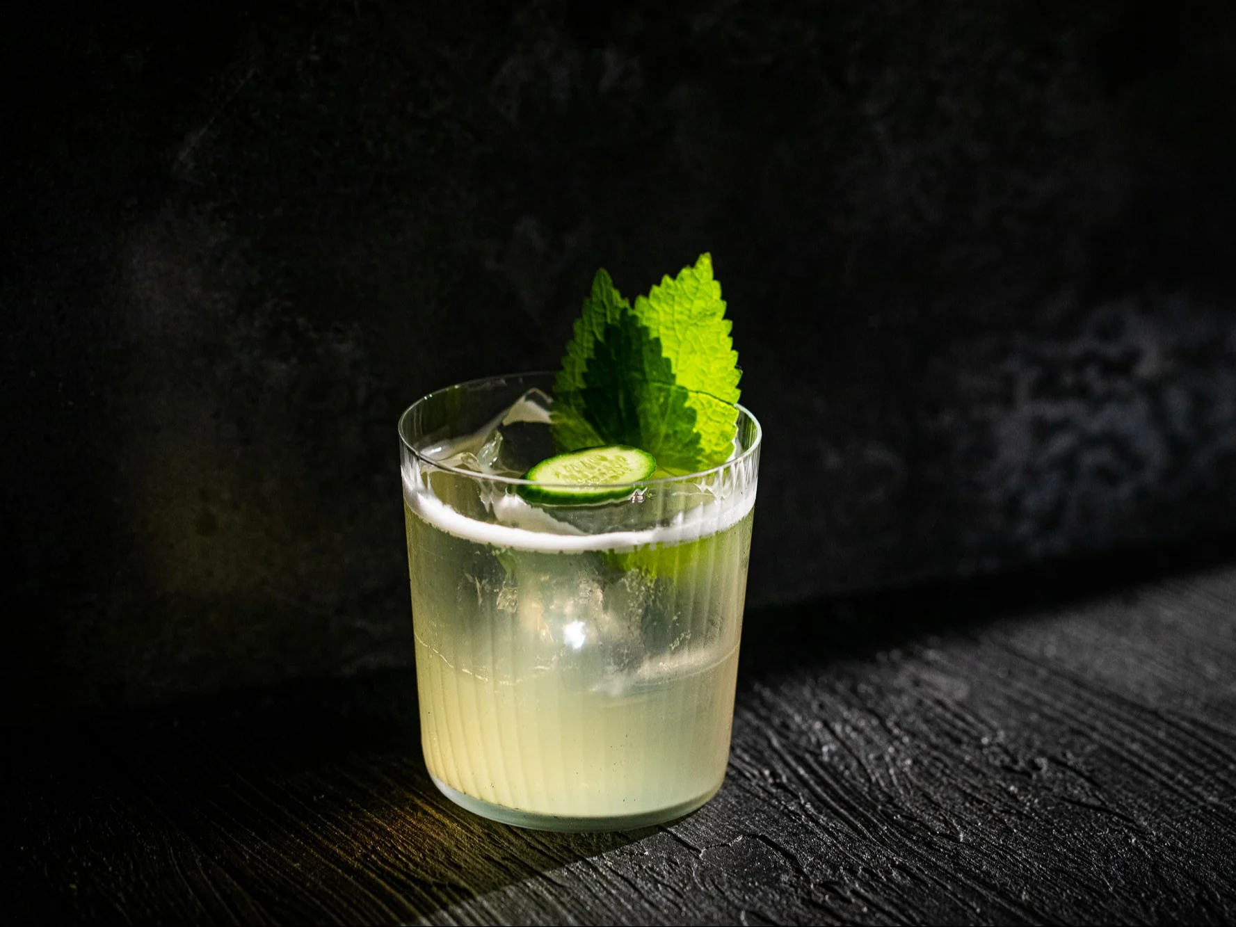 A pale cocktail in a rccks glass, with ice, a cucumber slice and two mint leaves