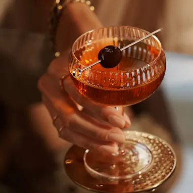 A Manhattan cocktail in a ridged coupe glass sits on a glass tables. It has a skewered maraschino cherry laying across the rim of the glass. A person holds the glass by its stem.