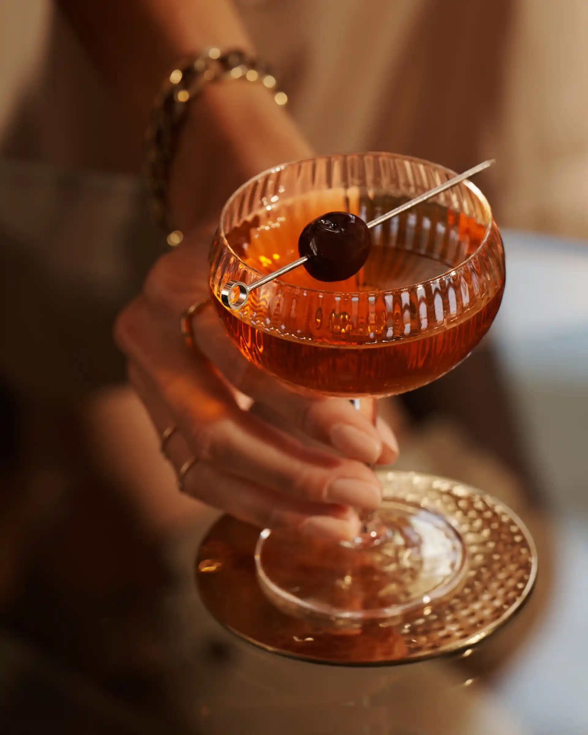 A Manhattan cocktail in a ridged coupe glass sits on a glass tables. It has a skewered maraschino cherry laying across the rim of the glass. A person holds the glass by its stem.