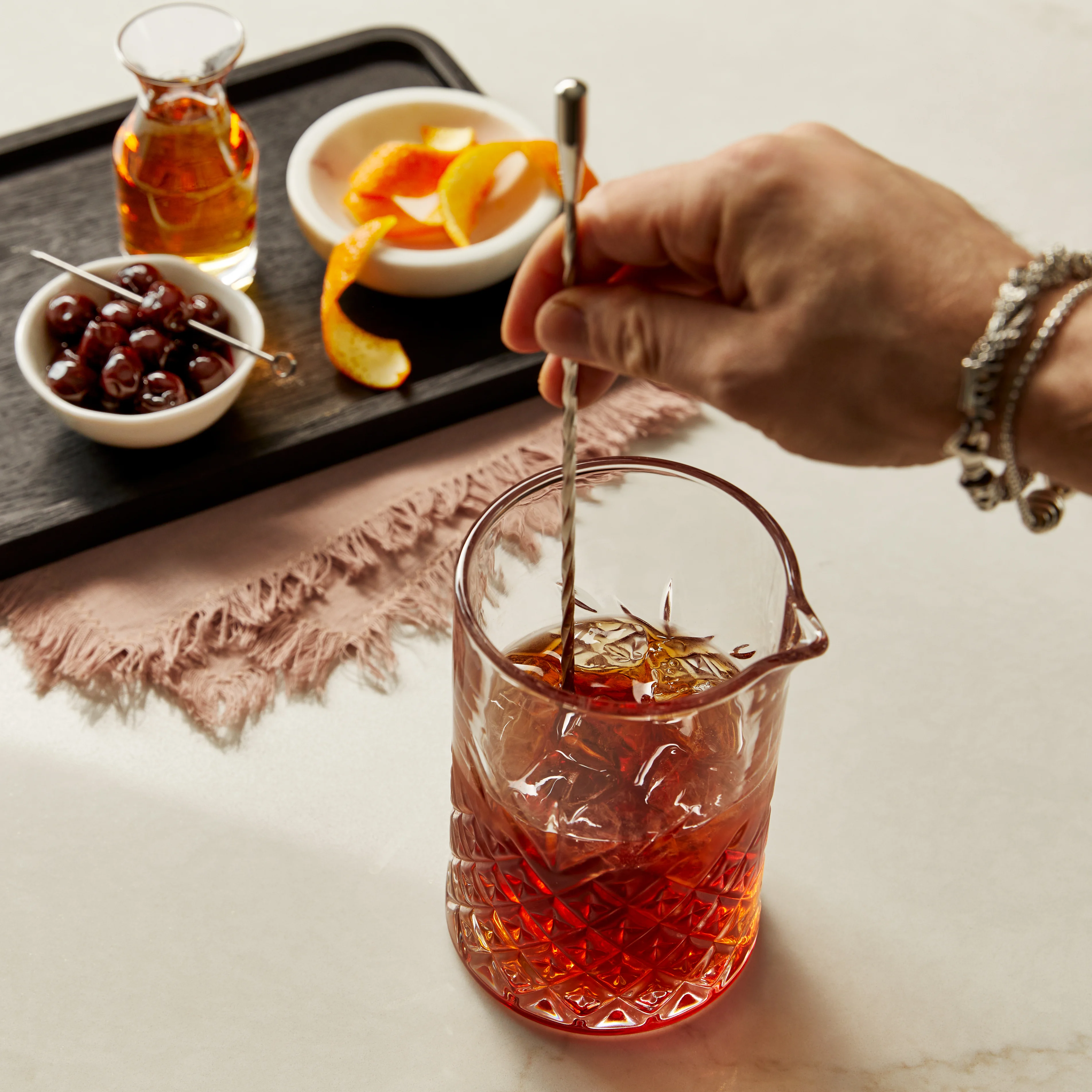 Een hand roert een lange metalen barlepel in een glas whiskycocktail, op de tafel ernaast staan een schaaltje olijven, een schaaltje sinaasappelgarnituren en een kleine glazen fles whisky.  