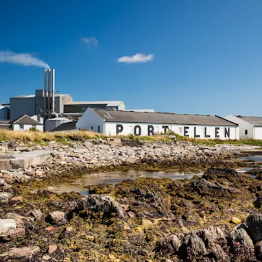 A while building is seen in the background, with the words Port Ellen painted in black in large letters. In front of the building are several rocks surrounding a body of water. The water is calm and the sky is bright blue with one cloud.