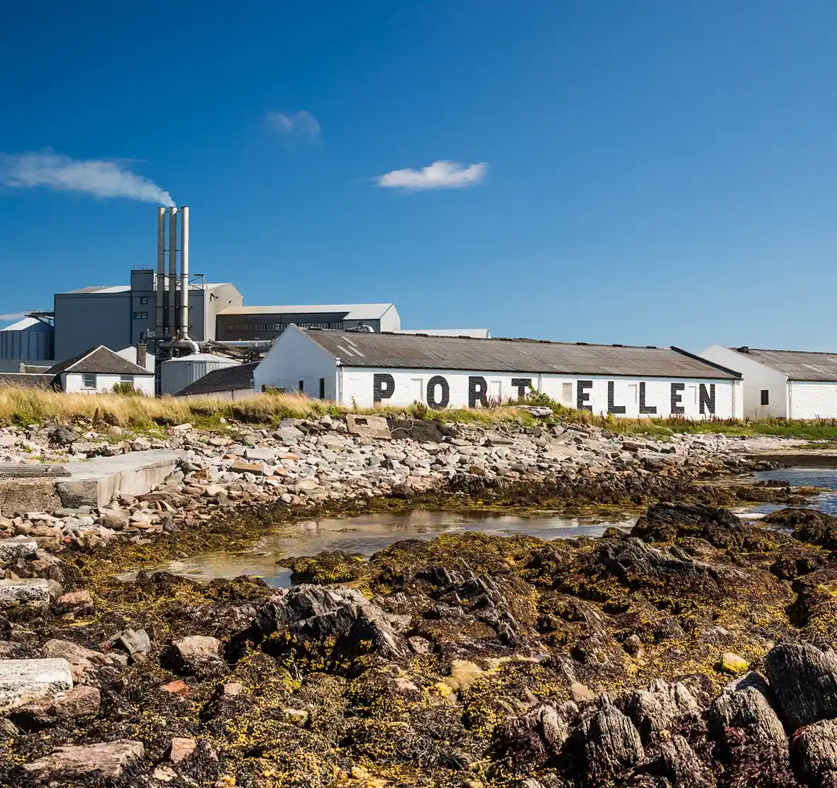 A while building is seen in the background, with the words Port Ellen painted in black in large letters. In front of the building are several rocks surrounding a body of water. The water is calm and the sky is bright blue with one cloud. 