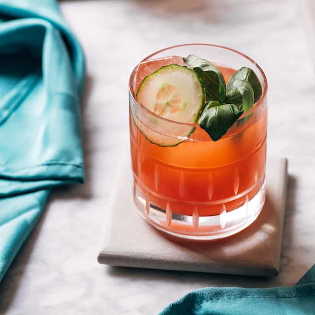 A pink cocktail in a rocks glass with ice and a cucumber garnish.