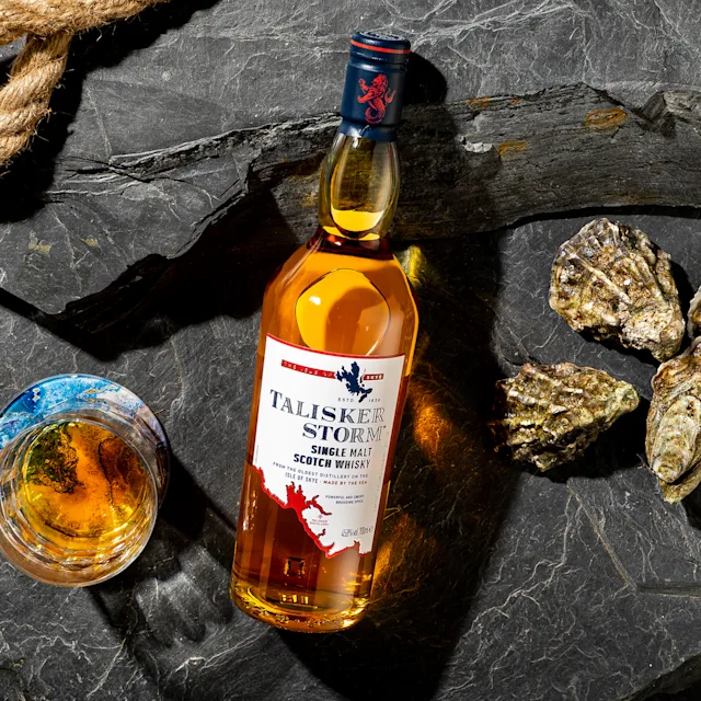 Kruidige whisky kopen Een fles Talisker Storm ligt op zijn rug op een leistenen ondergrond, omringd door gesloten mosselen en een rocks glas met whisky.