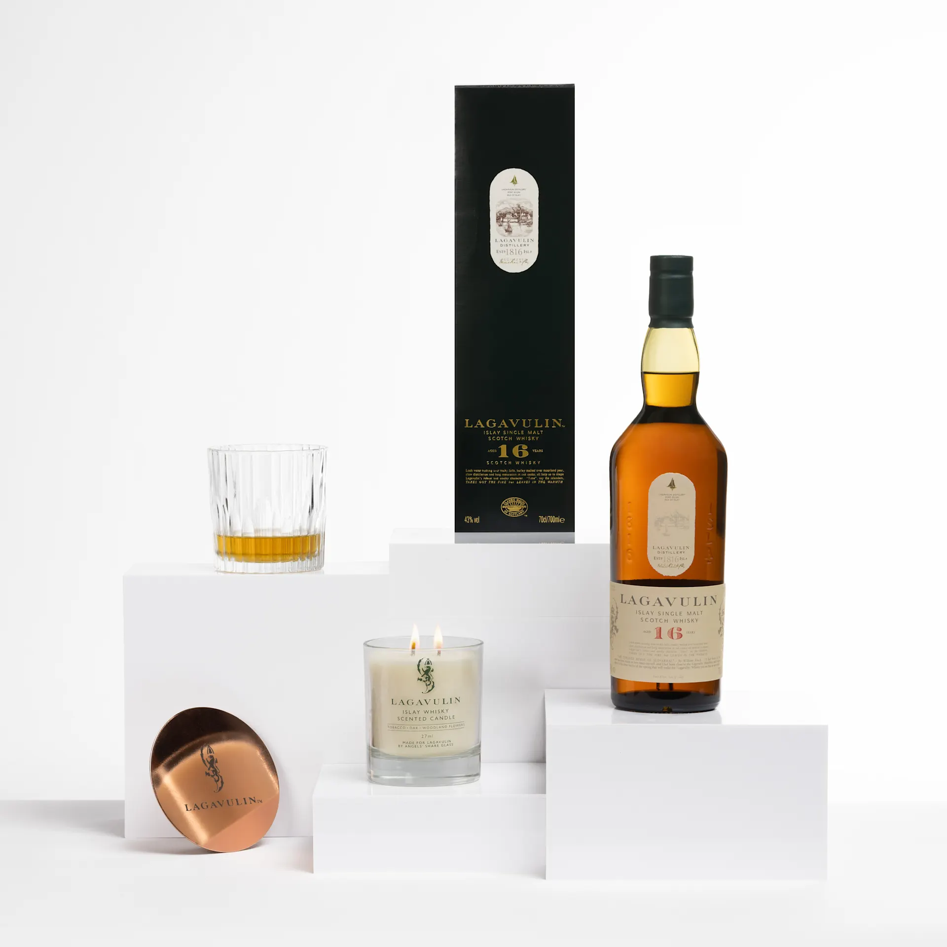 A bottle of Lagavulin 16 Year Old Scotch whisky, a branded candle with two wicks in a glass jar, and a copper-toned coaster with the Lagavulin logo, arranged on minimalist white blocks.