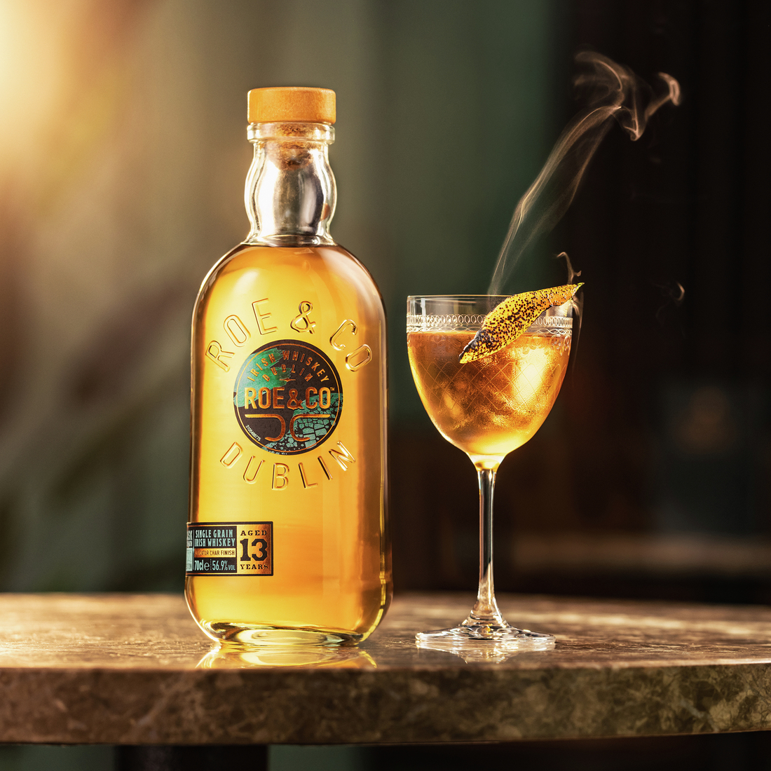 Roe & Co 13-Year-Old Cask Strength Irish Whiskey bottle next to a crafted cocktail garnished with a smoked citrus twist, on a marble bar top.