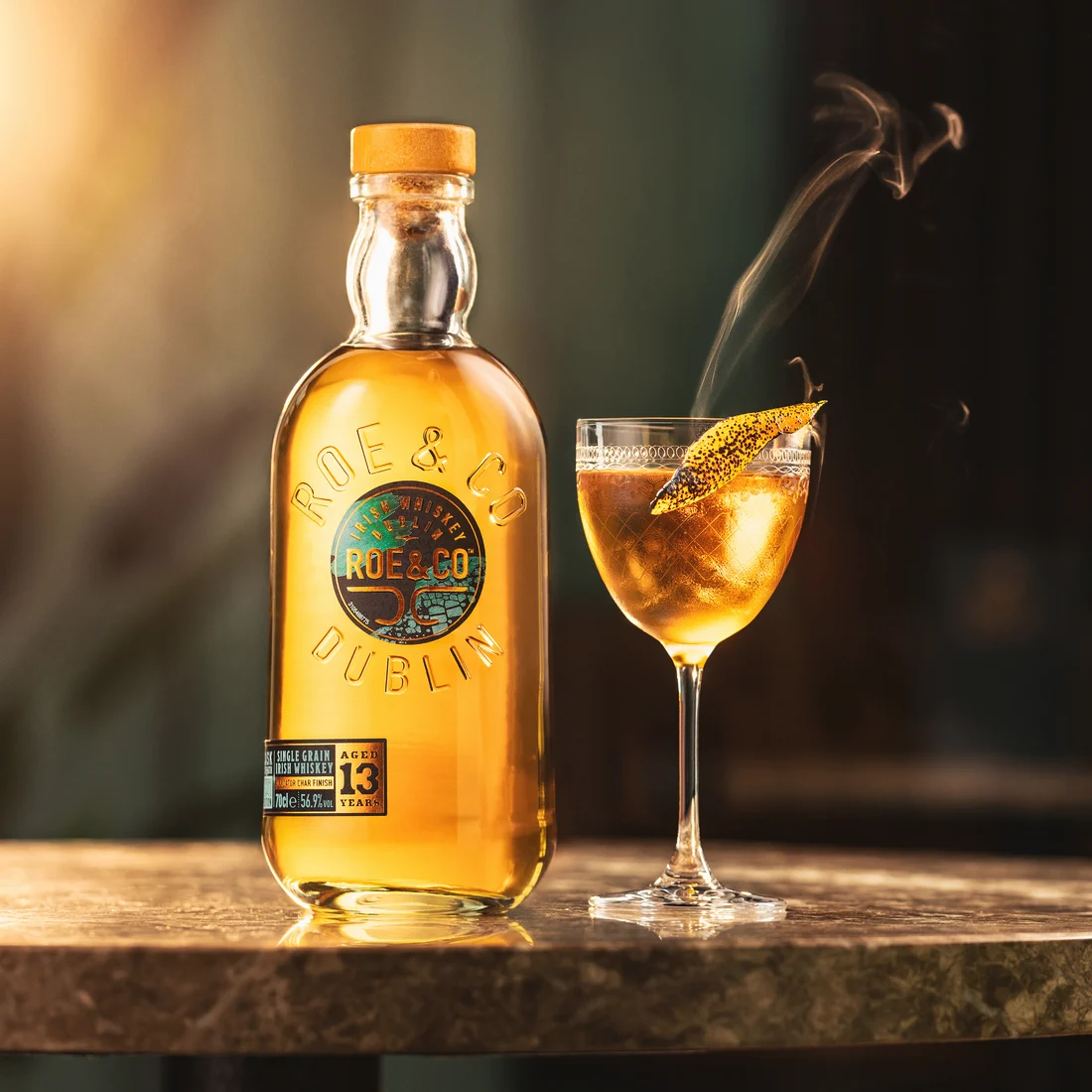 Eine Flasche Roe & Co 13-Year-Old Cask Strength Irish Whiskey neben einem selbstgemachten Cocktail, garniert mit einer geräucherten Zitrusfrucht, auf einer Marmorbar.