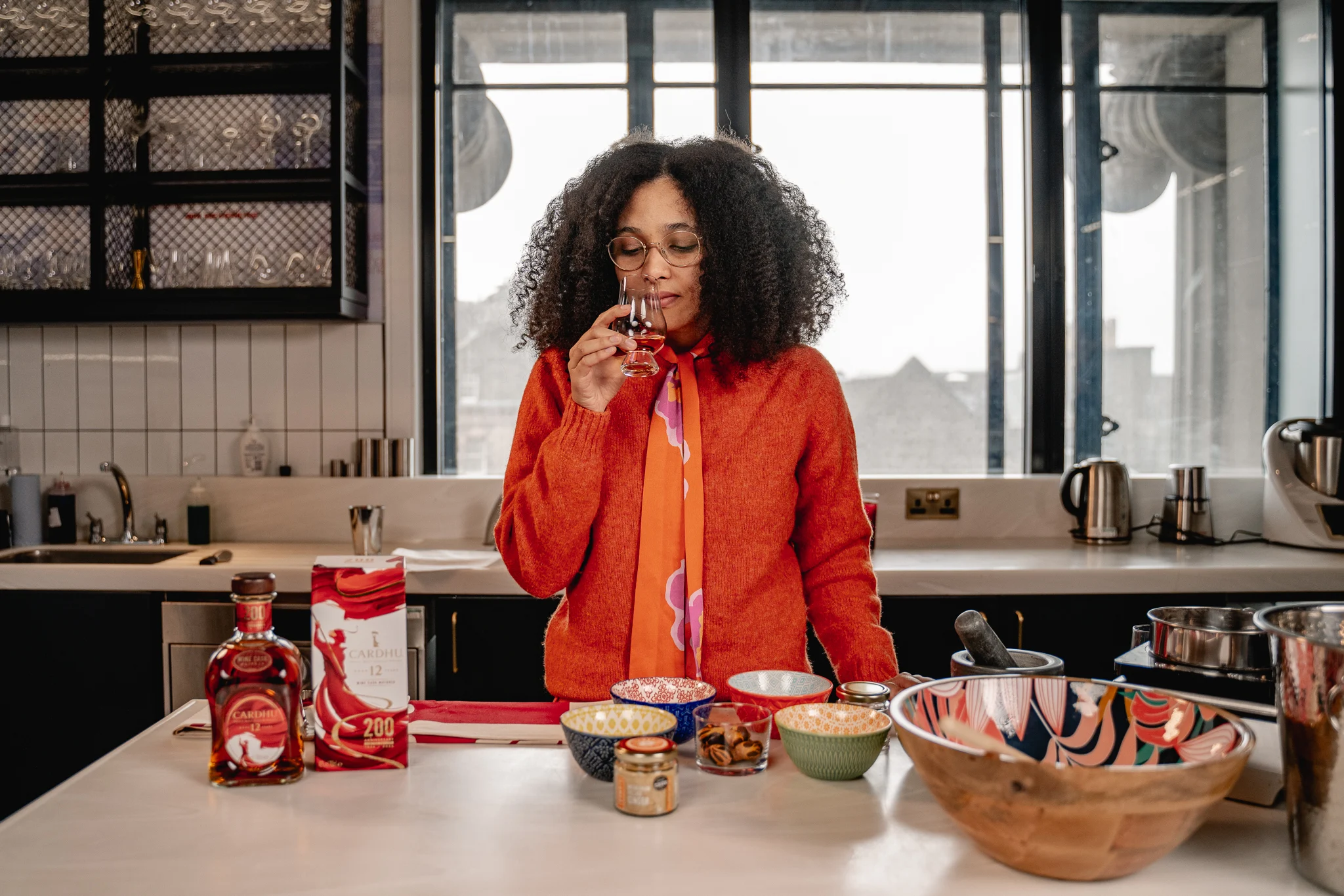 Jawahir stands in a well-lit kitchen surrounded by mixing bowls, ingredients, and a bottle of Cardhu Single Malt Scotch Whisky. Jawahir has their nose over a dram to smell the whisky.