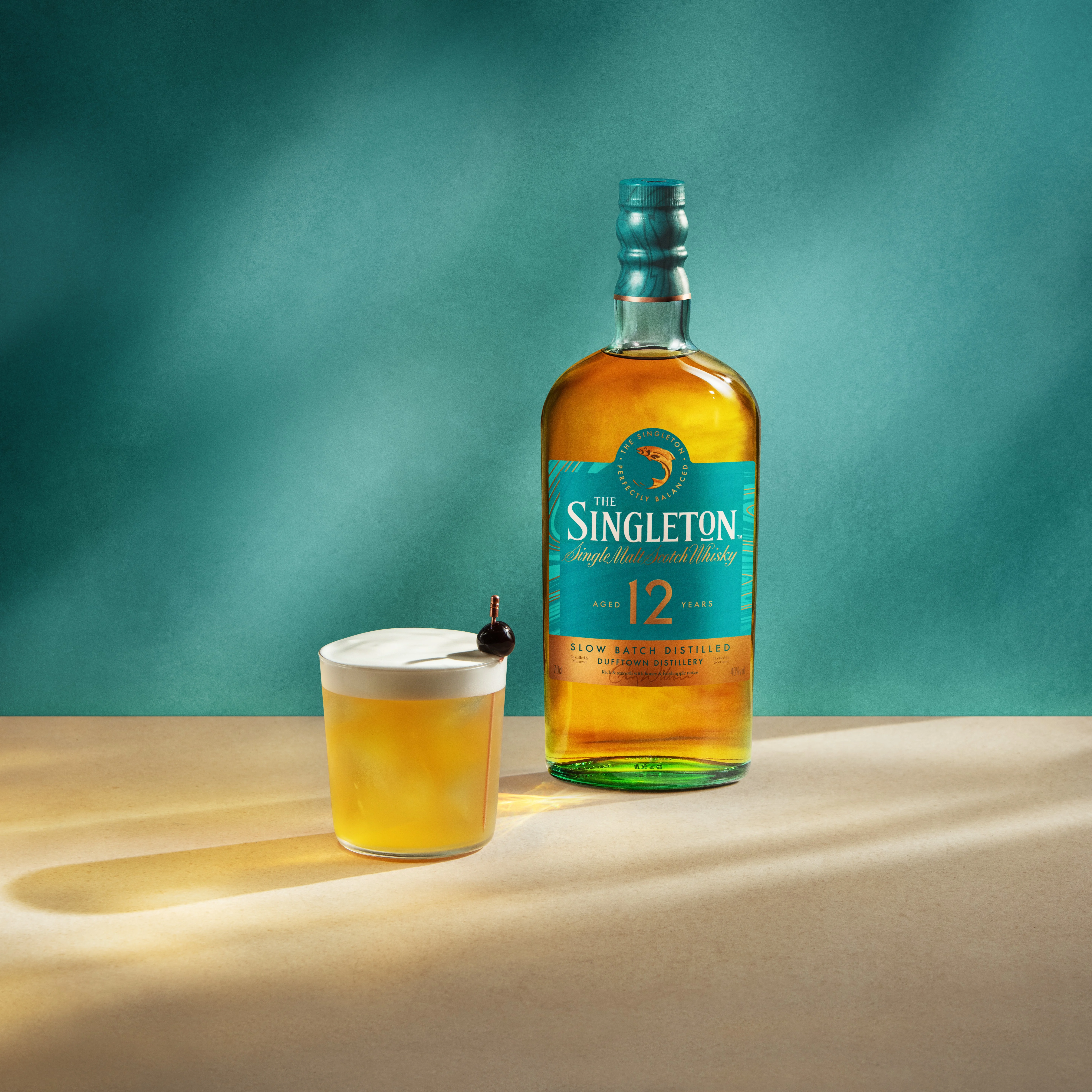 A bottle of The Singleton 12 Year Old Whisky sits next to a whisky sour cocktail in a rocks glass with a skewered maraschino cherry.