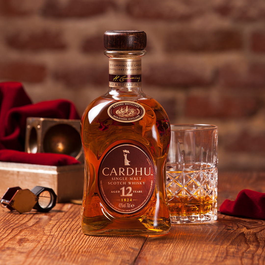 A bottle of Cardhu 12 year old whisky sits on a wooden table next to a rocks glass containing whisky