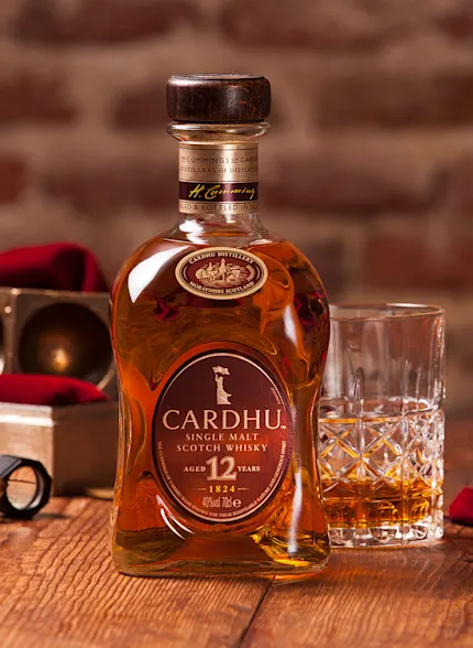 Eine Flasche Cardhu 12 Jahre alter Whisky steht auf einem Holztisch neben einem Steinglas mit Whisky