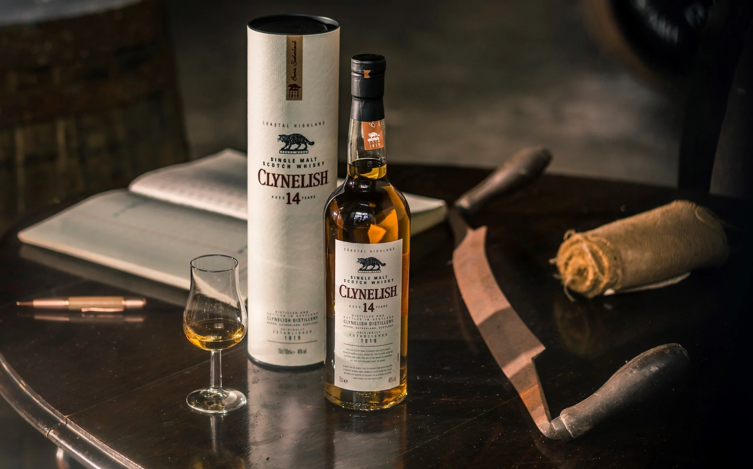 A bottle of Clynelish whisky sits on a circular wooden table, with its outer tube standing behind it. Also on the table is a dram glass containing whisky, a pen, notebook and a rolled-up napkin. Behind the table is a wooden barrel