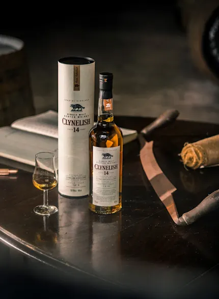 A bottle of Clynelish whisky sits on a circular wooden table, with its outer tube standing behind it. Also on the table is a dram glass containing whisky, a pen, notebook and a rolled-up napkin. Behind the table is a wooden barrel